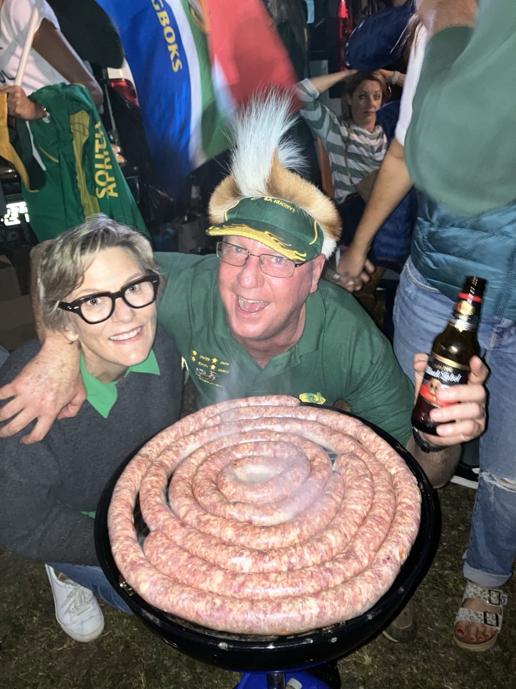 Two guests in Springbok green behind a boerewors coil on the braai at night