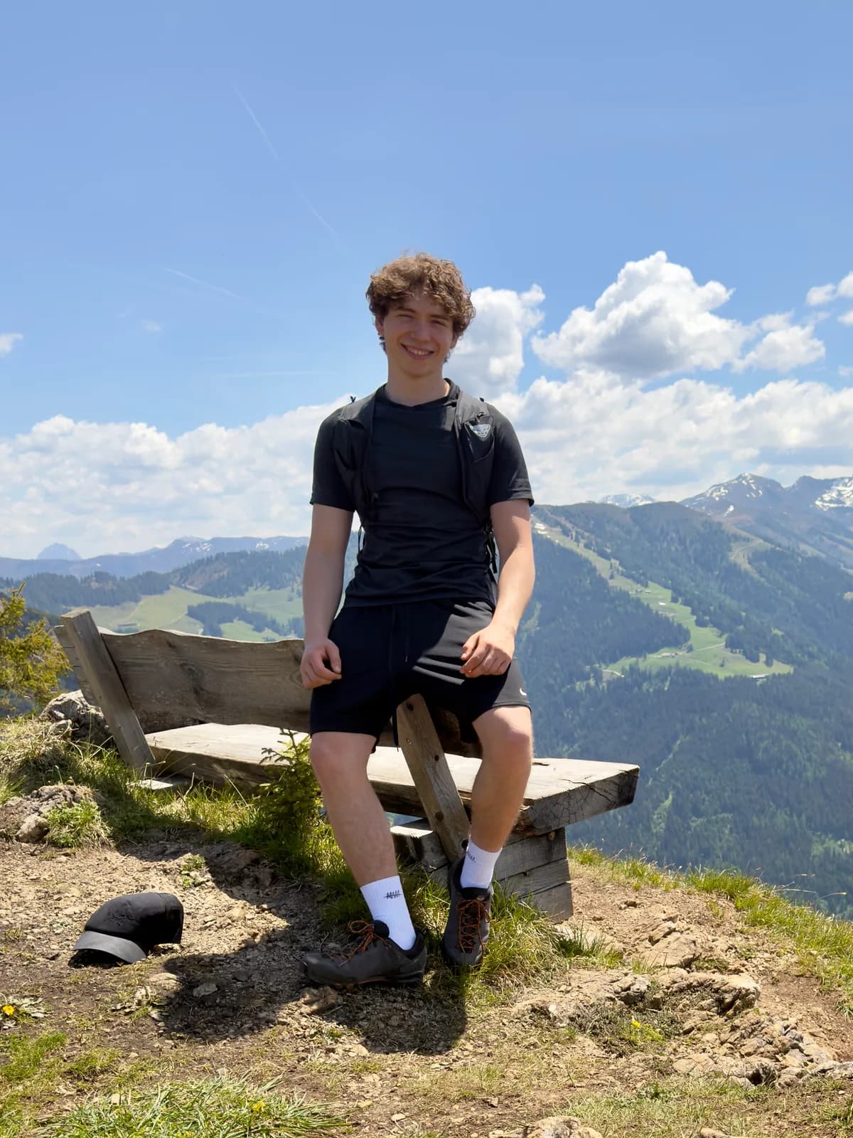 Fabio leaning against a weathered wooden bench on a Tirolean summit, green valley and snow-flecked peaks behind him
